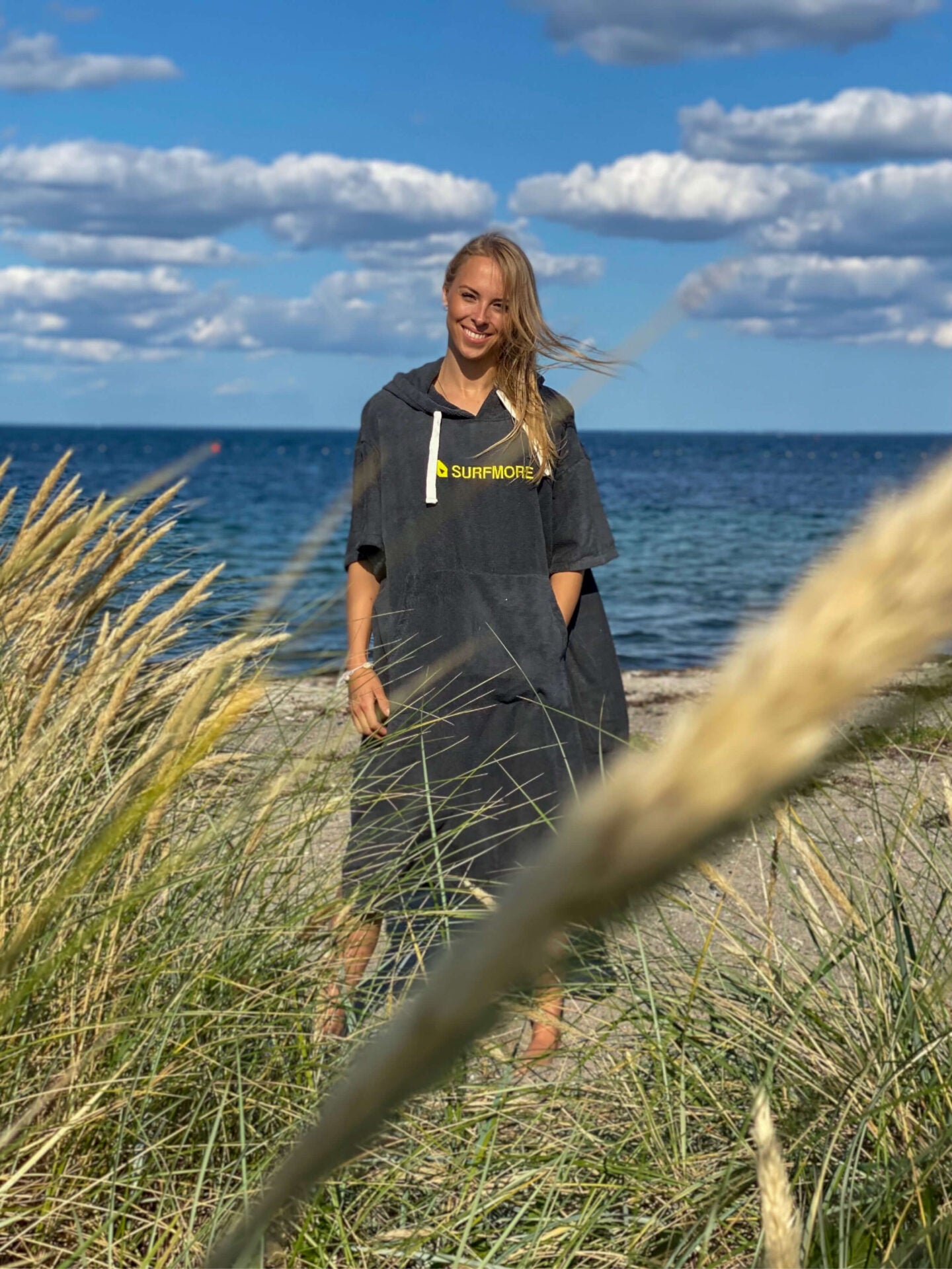 Ung og smilende kvinde iført en badeponcho i grå med hætte, snore og SURFMORE logo på maven